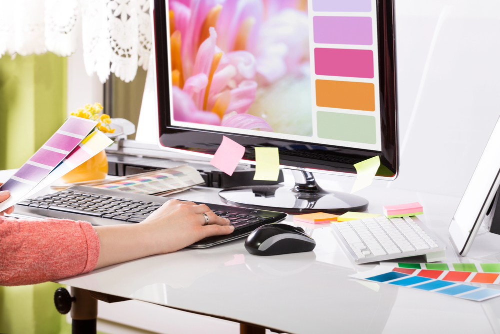 A desktop computer with a person's hand on the keyboard showing various color palettes on paper and one matching one onscreen